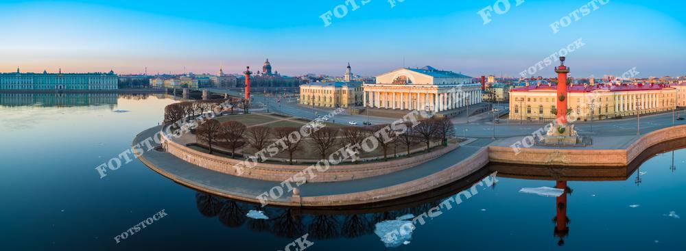 На фотографии изображена Стрелка Васильевского острова в Санкт-Петербурге в утренние часы. Ростральные колонны и здание Биржи отражаются в спокойной водной глади реки Невы. Нежные оттенки рассветного света добавляют картине романтичности и уюта. Вдали виднеется Исаакиевский собор, завершая панорамный вид исторического центра города.